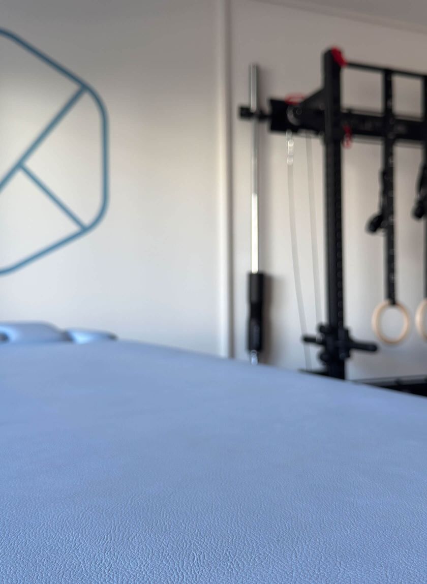 Blaue Behandlungsliege im Hintergrund eine Wand mit Fitnessgeräten für Physiotherapie.